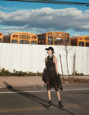 A woman in a floral dress and baseball cap stands on an asphalt road with parked yellow school buses in the background under a blue sky.