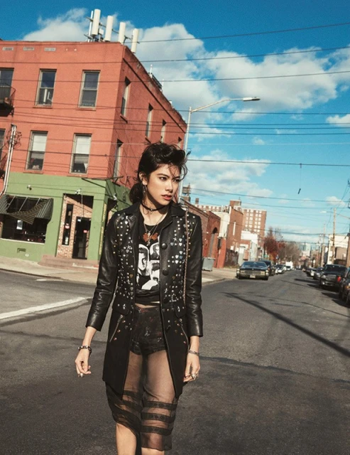 A young woman in a black studded jacket and sheer skirt walks down an urban street with brick buildings under a partly cloudy sky.
