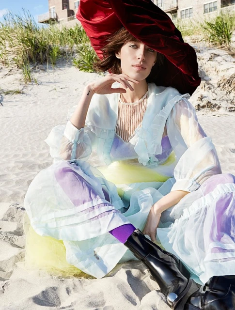 A woman poses on a beach wearing a pastel dress, black boots, and a large red hat in an outdoor fashion shot.