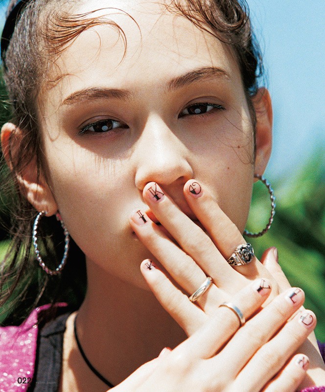 Close-up of a young woman with fair skin and hoop earrings. Her fingers are adorned with small black drawings in an outdoor setting.