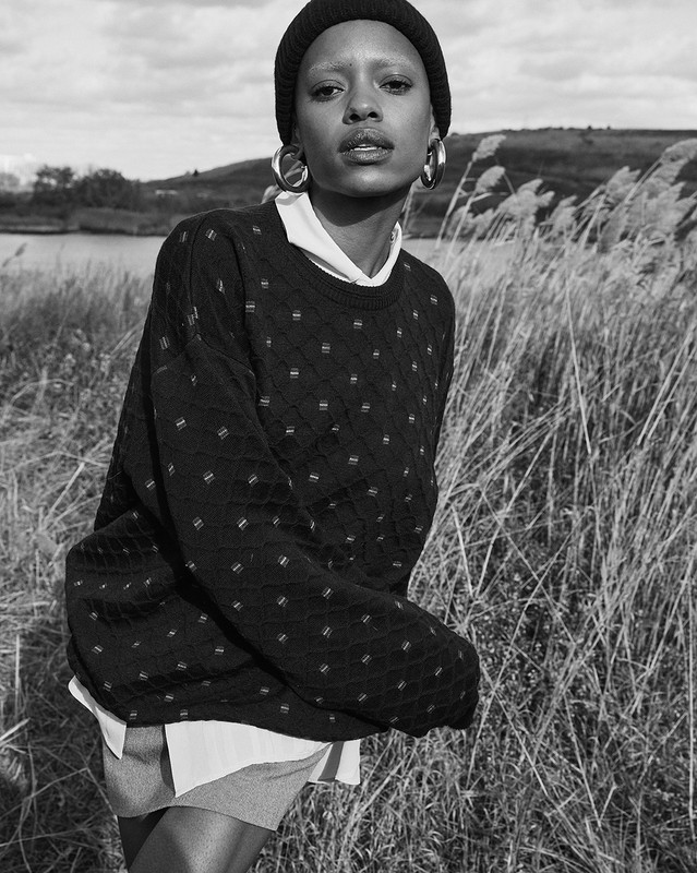 Monochrome portrait of a black woman in a knit beanie, oversize sweater, and hoop earrings standing in tall grass with a lake backdrop.