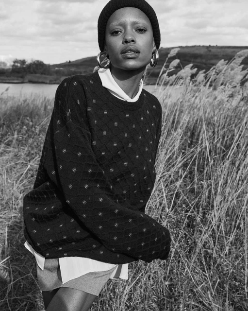 Monochrome portrait of a black woman in a knit beanie, oversize sweater, and hoop earrings standing in tall grass with a lake backdrop.