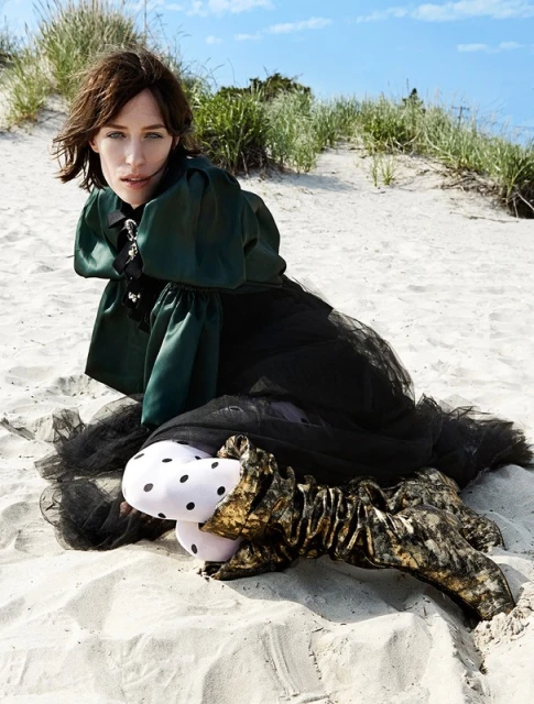 A brunette adult woman wearing a dark dress with green puffy sleeves and unique patterned stockings poses on a sandy beach under a bright sky.