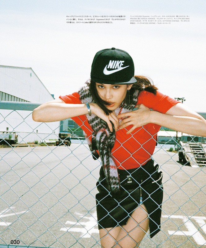 A young woman in a black Nike cap, red t-shirt, and black skirt leans against a wire fence outdoors on a sunny day.