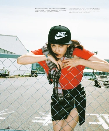 A young woman in a black Nike cap, red t-shirt, and black skirt leans against a wire fence outdoors on a sunny day.