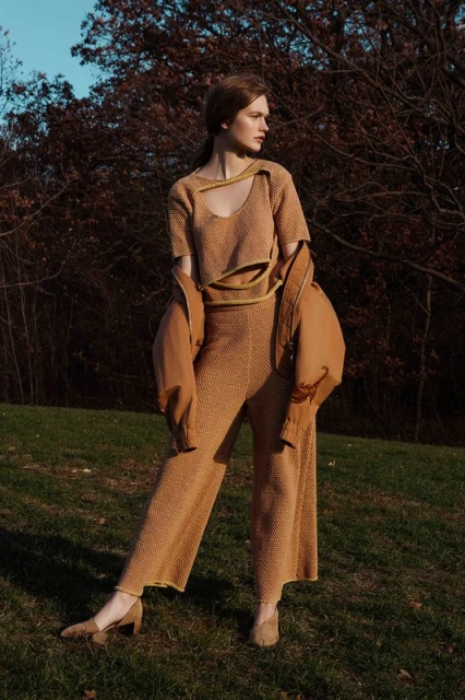 A woman wearing a brown knitted matching set poses outdoors, jacket draped over her shoulder, with a forest background on a sunny day.