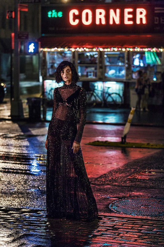 Fashionable woman in a sheer, dark sparkly dress poses on a wet city street corner at night under bright neon signage.