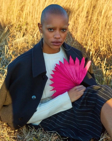Close-up portrait of a bald woman with bleached eyebrows holding a bright pink folded fan in dry tall grass.