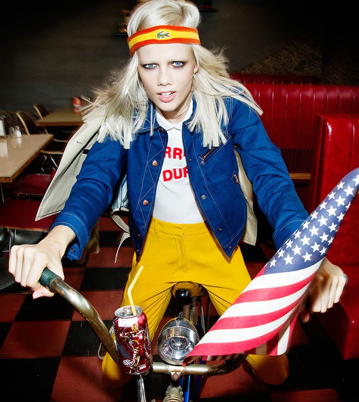 A fashionable woman rides a bike indoors with a can and flag attached, wearing a blue jacket, yellow pants, and a headband.