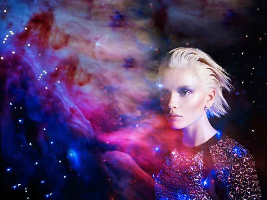 Woman with platinum blonde hair and makeup stands in front of nebula background wearing a sparkling dark blouse in portrait format.