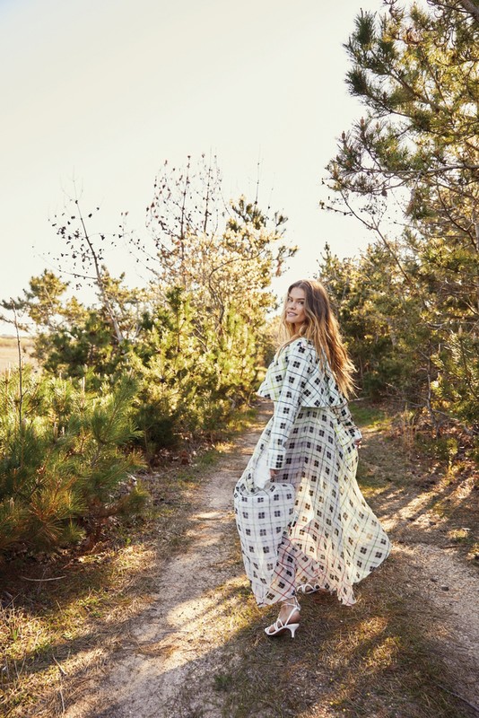 Woman in a long patterned plaid dress walking along a dirt path through a pine forest with sunlight filtering through the trees.