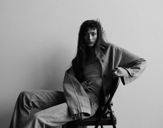 Monochrome portrait of a woman in denim clothing seated on a chair against a plain wall.