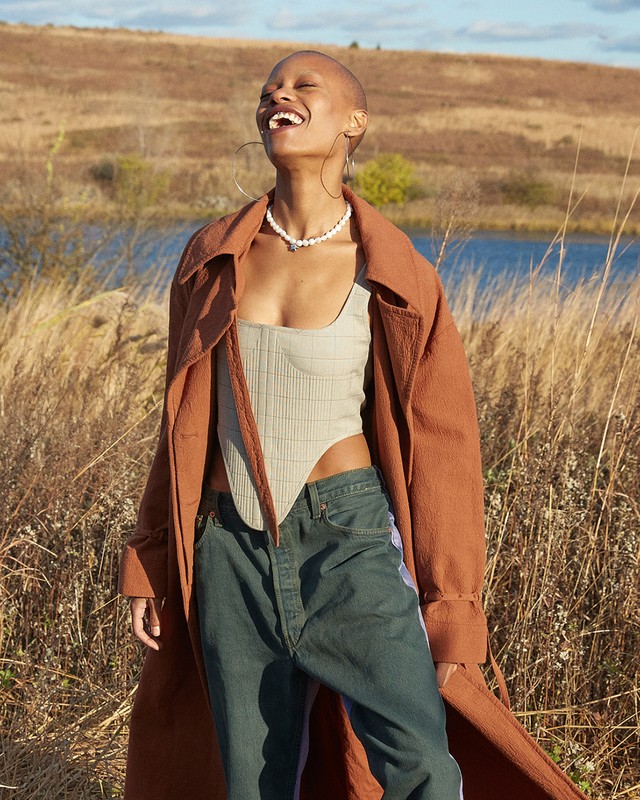 Portrait of an adult woman laughing in a rural field near a lake, wearing a brown coat and denim jeans, with a bright sunny sky.