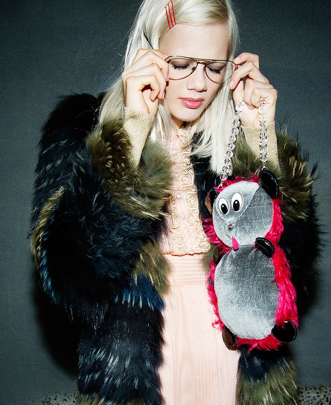 A blonde woman in a furry coat adjusts her glasses while holding an owl-shaped purse against a muted gray backdrop in a studio shot.