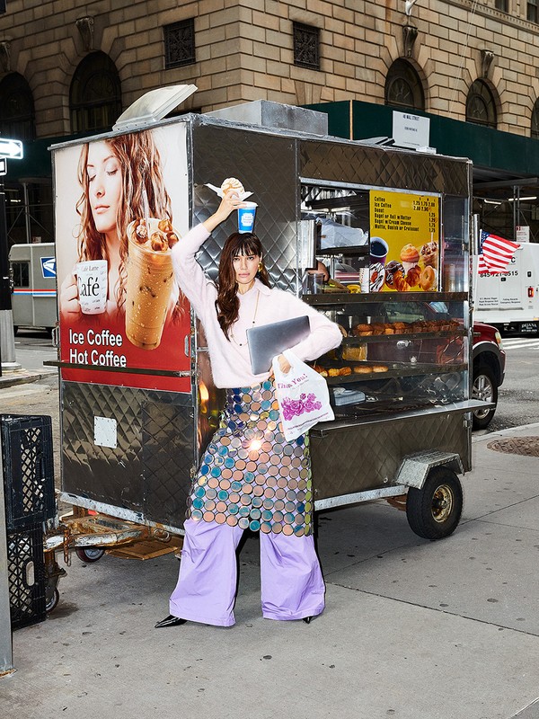 A woman in fashionable clothing poses in front of a food cart in an urban setting, holding a drink and laptop, with passersby in the background.