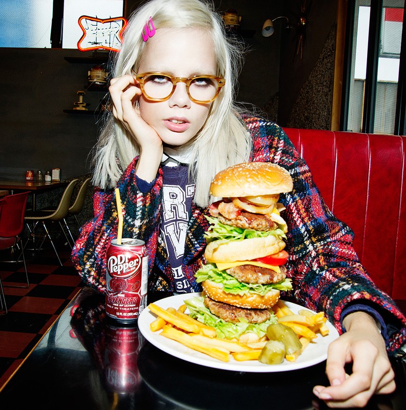 Young woman wearing glasses poses with a large hamburger, fries, and a Dr. Pepper in an indoor restaurant setting with red booths.