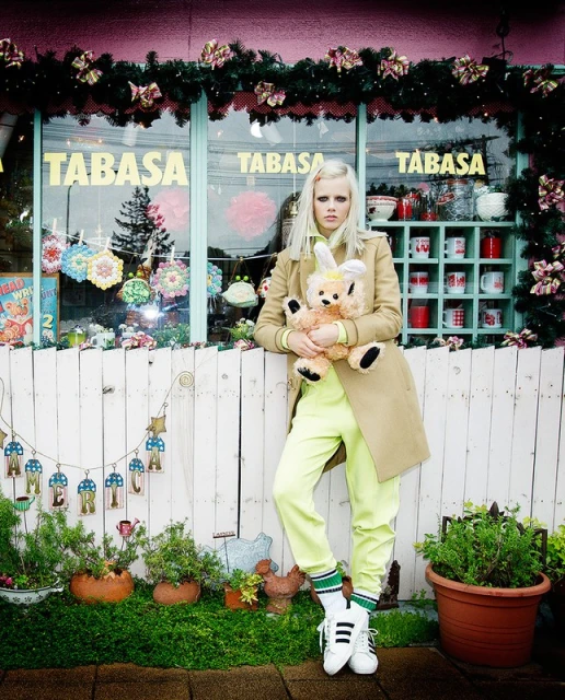 Woman with blond hair in a beige coat holding a teddy bear standing in front of a store with Christmas garlands and decorations.