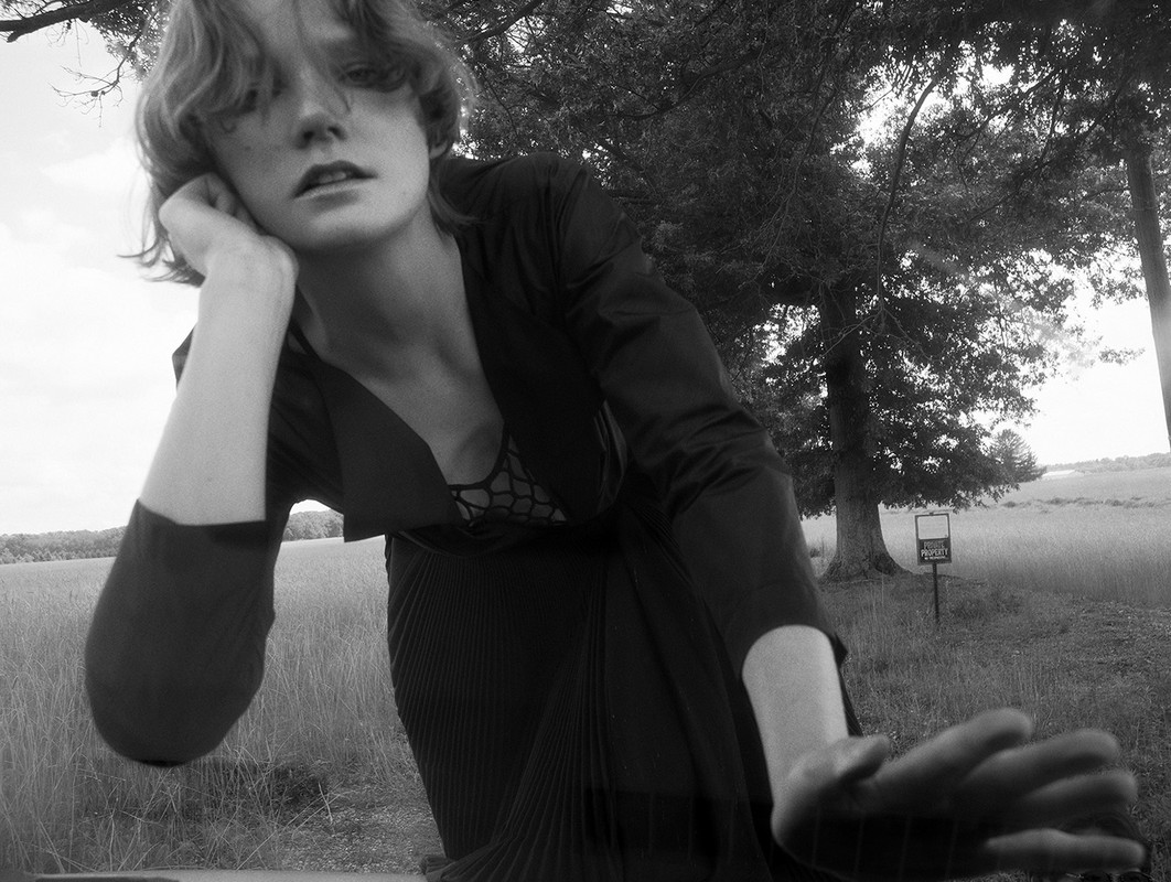 Black and white portrait of a fair-skinned woman with short hair, wearing a jacket, and posing in a field with trees in the background.