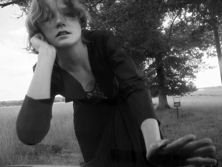 Black and white portrait of a fair-skinned woman with short hair, wearing a jacket, and posing in a field with trees in the background.