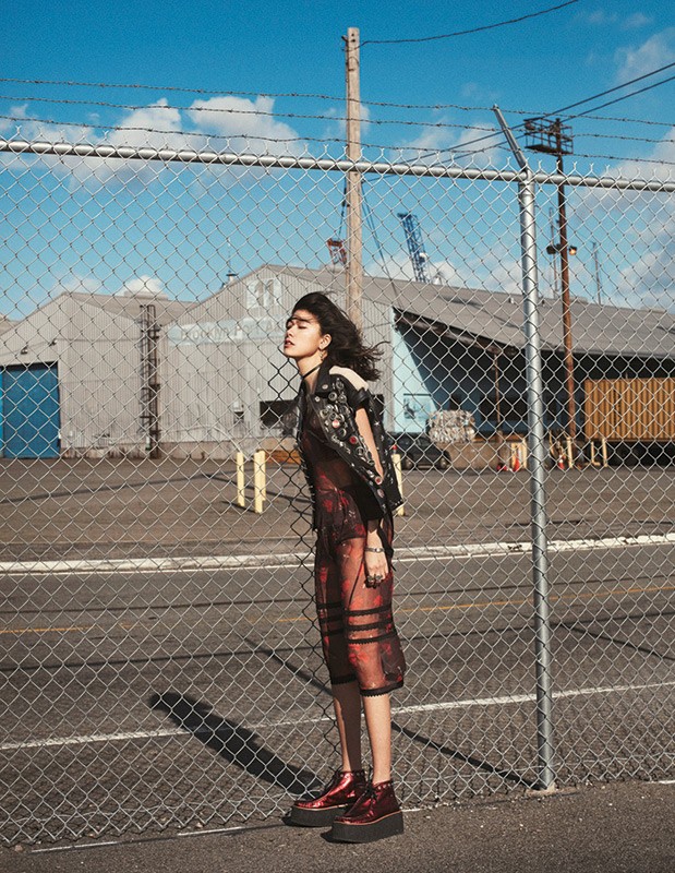 A young woman stands in a patterned dress and jacket with red platform shoes in front of a chainlink fence in an industrial outdoor setting.