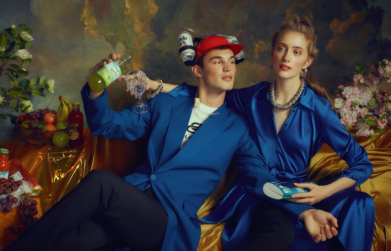 Elegant couple in studio with flowers and food, wearing stylish blue outfits and jewelry, set against a renaissance-style backdrop.