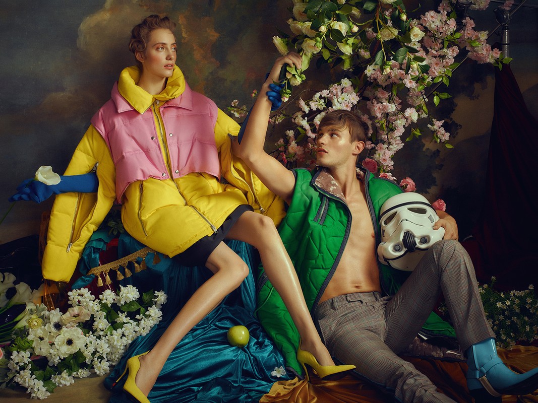Two fashion models strike a pose indoors, one holding a helmet, amidst flowers and lush fabrics, under studio lighting, editorial style.