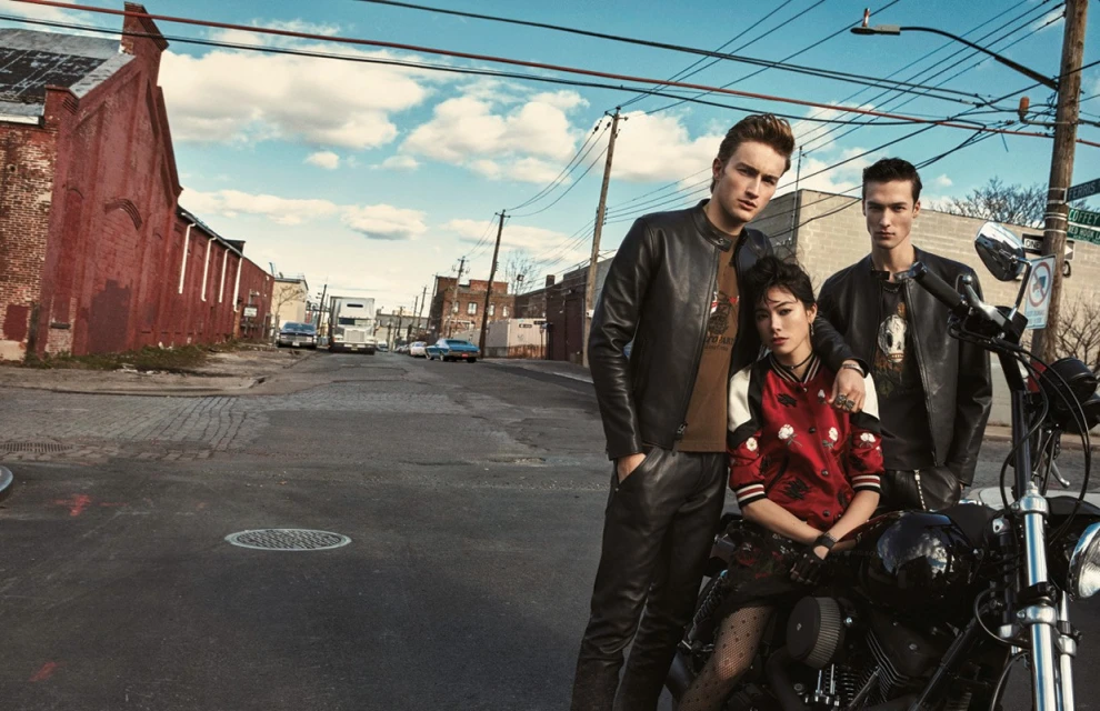 Group shot of two men and one woman posing with a motorcycle on an urban street with brick buildings, power lines, and cars on a cloudy day.