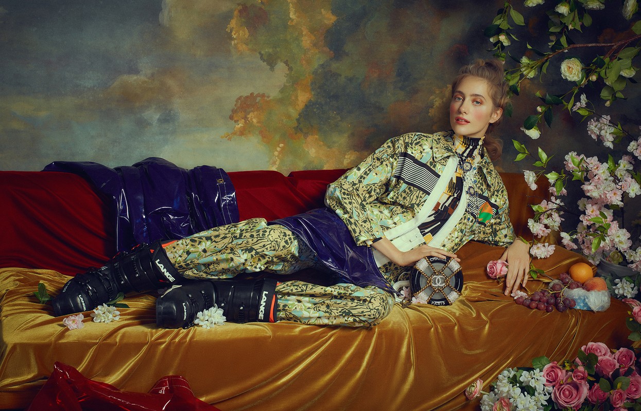 A fair-skinned woman poses on a plush, ochre couch surrounded by flowers, wearing a patterned jacket and ski boots, with a painted cloud backdrop.