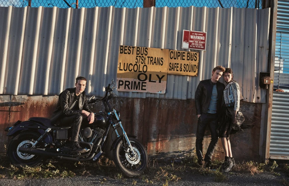 Two models pose with a black motorcycle against a weathered metal wall with vintage signage, exuding a raw, urban fashion aesthetic.