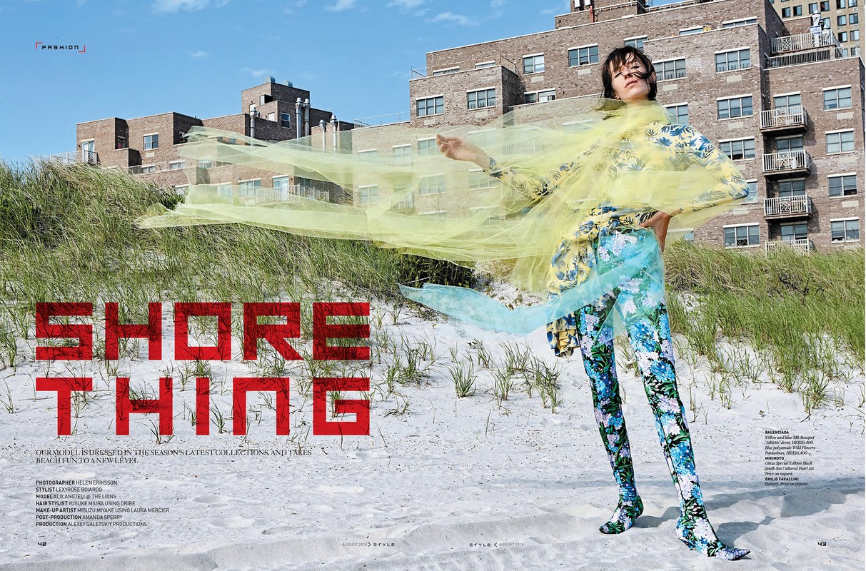 A fashion shot featuring a woman in a floral outfit and yellow mesh shawl on a beach, with high-rise buildings in the background, captured on a sunny day.