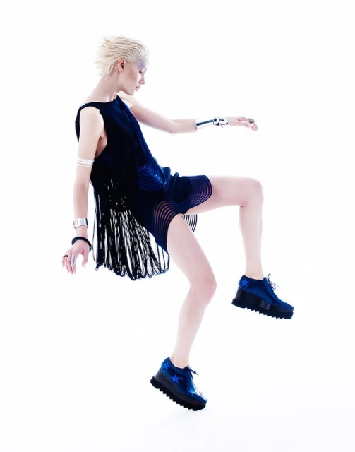 A woman with short blonde hair is dynamically posed mid-air in a dark dress and blue shoes against a bright white backdrop.