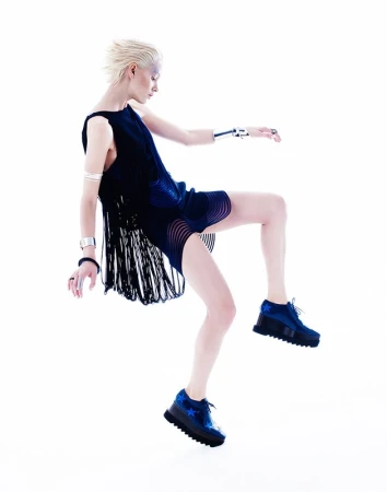A woman with short blonde hair is dynamically posed mid-air in a dark dress and blue shoes against a bright white backdrop.