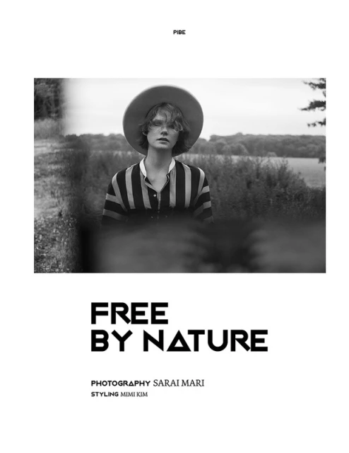 Black and white portrait of a young adult wearing a hat and striped shirt outdoors, surrounded by fields and natural vegetation.