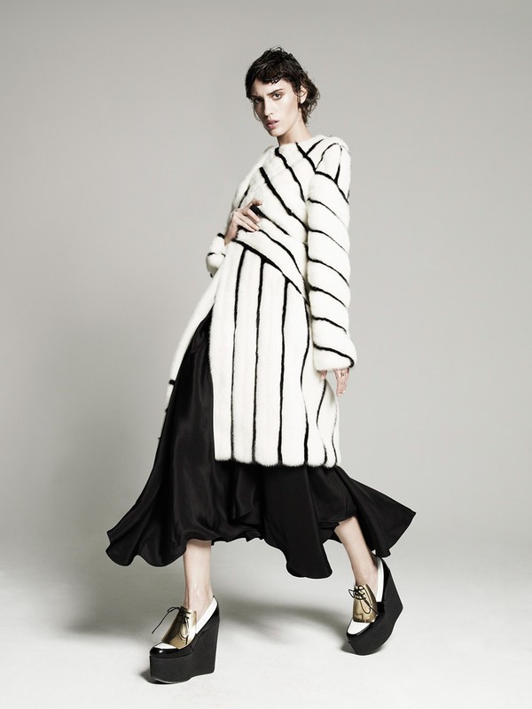 A fashionably dressed adult female model poses in a studio wearing a striped faux fur coat, black dress, and wedge platform shoes.