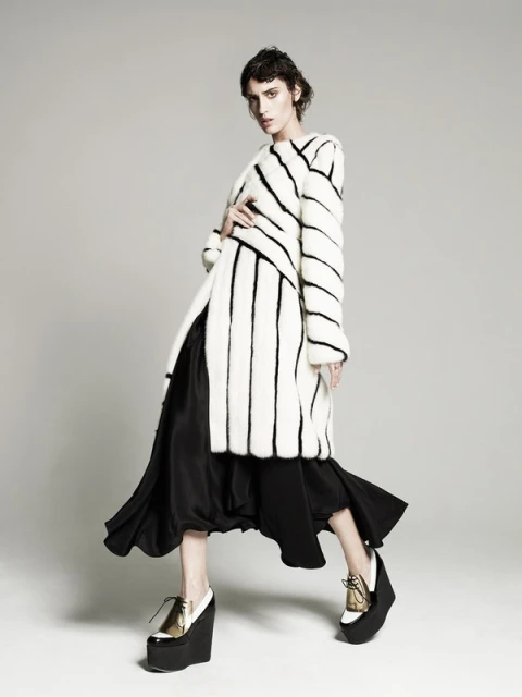 A fashionably dressed adult female model poses in a studio wearing a striped faux fur coat, black dress, and wedge platform shoes.