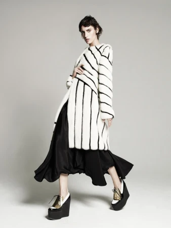 A fashionably dressed adult female model poses in a studio wearing a striped faux fur coat, black dress, and wedge platform shoes.