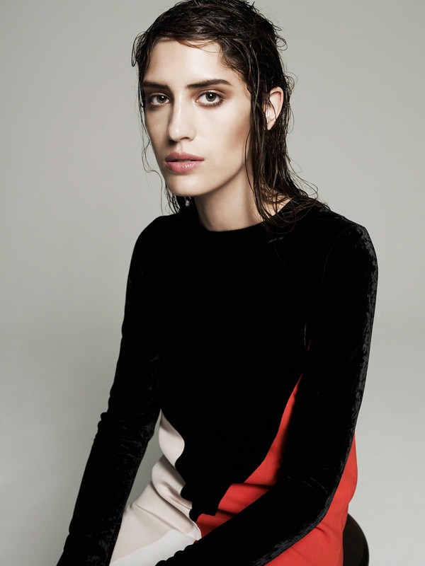 Portrait of a young adult woman with wet dark hair, wearing a color-block top with black, white, and red sections, set against a grey background.