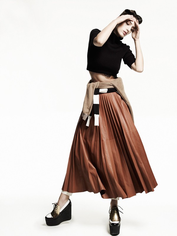 Elegant adult female fashion model poses in a black turtleneck crop top and long copper-colored pleated skirt, wearing platform shoes.