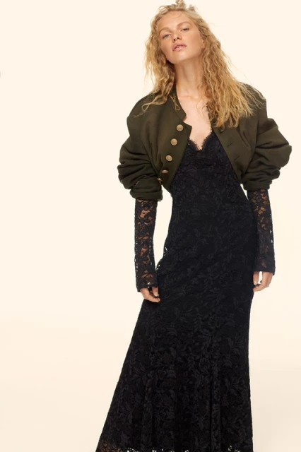 A blonde woman stands in a photo studio, wearing a black lace dress and a cropped olive-green jacket with gold buttons.