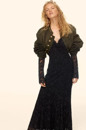A blonde woman stands in a photo studio, wearing a black lace dress and a cropped olive-green jacket with gold buttons.