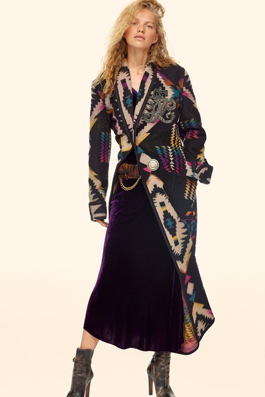 A young adult female with curly blonde hair is modeling an Aztec-print coat with a purple velvet dress and platform heel boots.