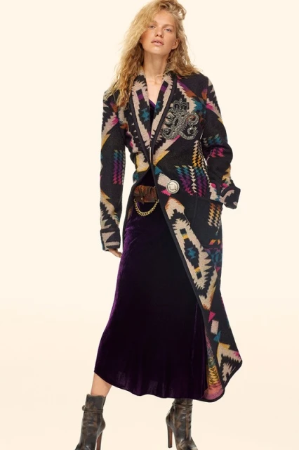A young adult female with curly blonde hair is modeling an Aztec-print coat with a purple velvet dress and platform heel boots.