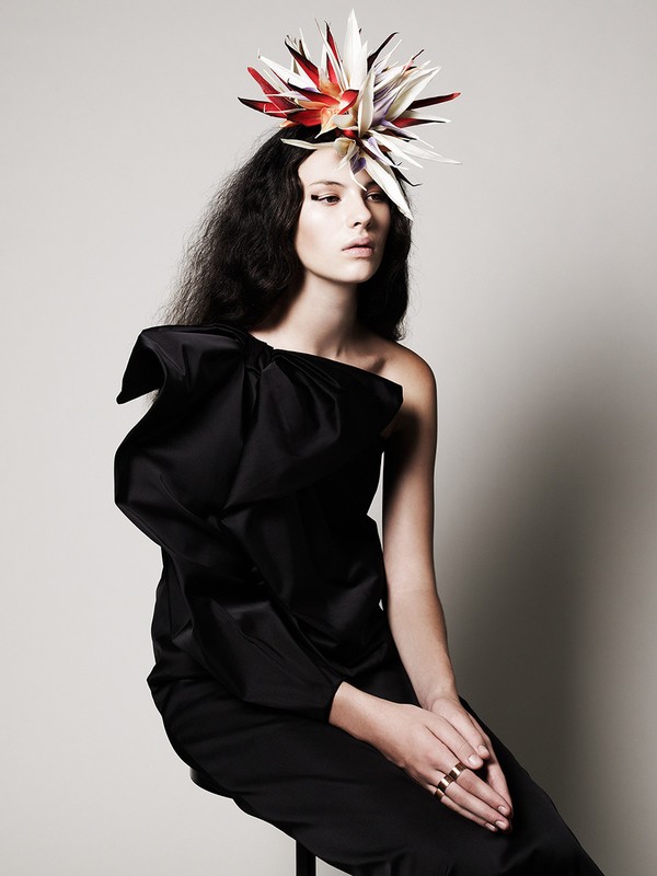 Elegant woman in a black one-shoulder dress poses gracefully with a unique floral headpiece against a soft grey studio backdrop.