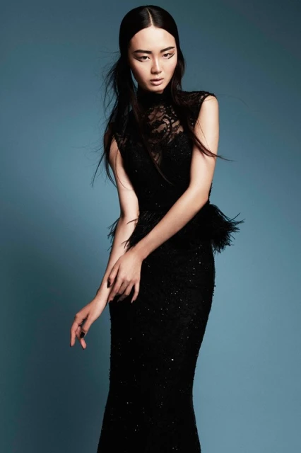 Elegant Asian woman in a black lace gown with feather detail posing against a blue background in a studio fashion photograph.