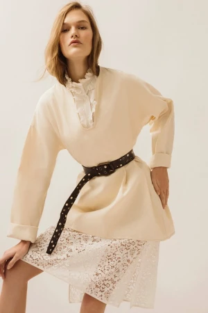 Model in cream blouse with ruffles and a black studded belt posing against a white studio background.