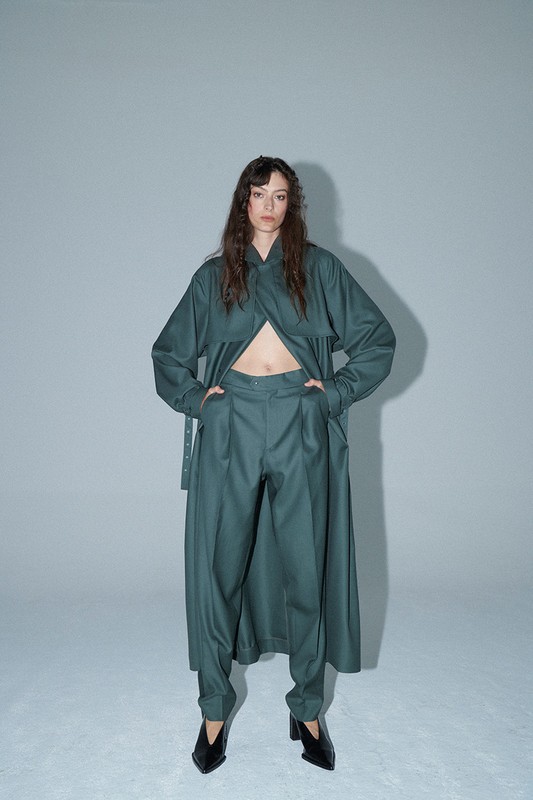 Full-length studio shot of a woman in a long-sleeved green jacket and green pants, looking at the camera against a grey background.