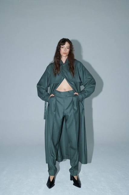 Full-length studio shot of a woman in a long-sleeved green jacket and green pants, looking at the camera against a grey background.