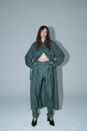 Full-length studio shot of a woman in a long-sleeved green jacket and green pants, looking at the camera against a grey background.