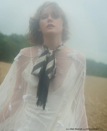 A woman stands in a misty field, wearing a white dress and a striped black-and-white scarf, creating an ethereal and dreamlike atmosphere.