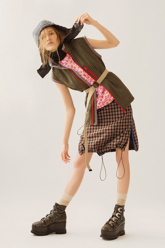 A woman wearing a gray bonnet, green vest, pink floral top, and plaid skirt strikes a pose in a fashion-forward shot in a studio setting.